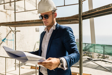 Man architect wearing formal suit and hard hat during building construction control holding a blueprints on a construction site