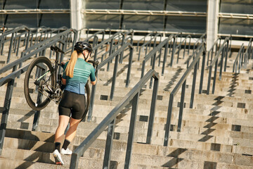 Full length shot of professional female cyclist in cycling garment and protective gear with bicycle on her shoulder walking up steps, training outdoors