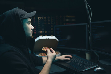 A hacker organize an attack on data servers. He hideout is dark and full of operational displays. The guy is taking a break, eating pizza and analyzing the malware code on the computer.