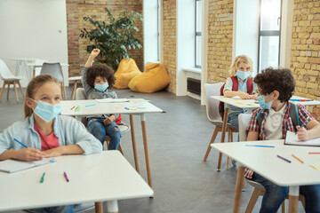 Obraz premium Little boy wearing protective face mask during coronavirus outbreak raised his hand in class. School kids studying in elementary school, sitting at the desk