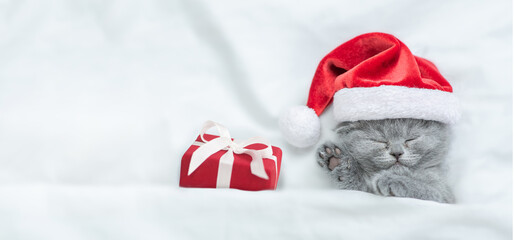 Cute kitten wearing red santa's hat sleeps with gift box under a white blanket on a bed. Top down...