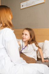 Fototapeta premium Back view of happy little girl listening heartbeat with stethoscope to female doctor sitting in bed. Cute child playing as doctor nurse listening physician with stethoscope. Concept of children health