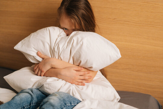 Upset Little Girl Crying Sitting Alone In Fetal Position Hiding Face And Rocking On Bed Hugging Big Pillow And Looking At Camera . Concept Of Child Depression.