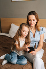 Happy young mother hugging to preschool daughter together browsing and surfing internet on mobile phone, chatting on video call sitting on bed in bedroom. Concept of family leisure activity at home.