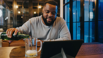 Middle-aged African American male drinking beer having fun happy night party event online celebration via video call in living room at home. Social distancing, quarantine for coronavirus prevention.