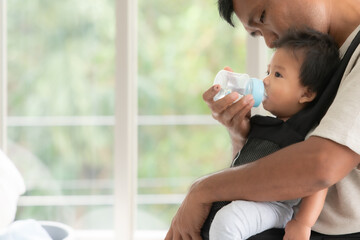 Asian father holding and feeding her baby boy with milk from baby bottle.