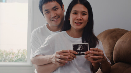 Young Asian Pregnant couple show and looking ultrasound photo baby in belly. Mom and Dad feeling happy smiling peaceful while take care child lying on sofa in living room at home concept.