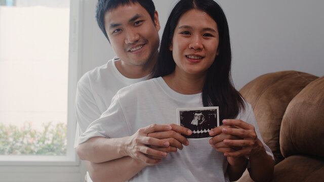 Young Asian Pregnant couple show and looking ultrasound photo baby in belly. Mom and Dad feeling happy smiling peaceful while take care child lying on sofa in living room at home concept.