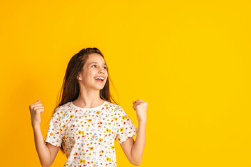 cheerful girl of about 10 years old in a floral T-shirt with her hands clenched into fists and a happy smile on a yellow background. A happy child enjoys success and victory. Discounts and sales