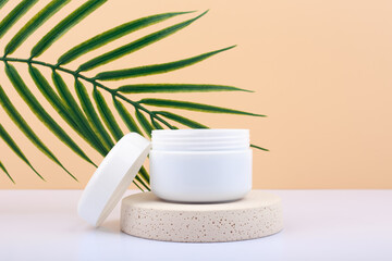 White opened cosmetic jar on beige podium against beige background with palm leaf. Concept of cosmetic products for daily skin or hair care. Jar with scrub, mask or balm.