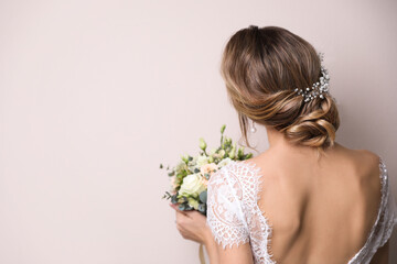 Young bride with elegant hairstyle holding wedding bouquet on beige background, back view. Space...