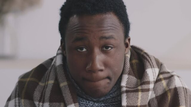 Close up portrait of sick african american guy wrapped in plaid drinking healing tea and looking sadly at camera