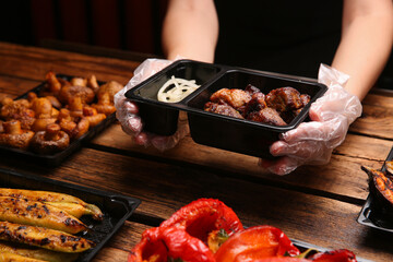 Waiter with plastic container of tasty shish kebab at wooden table, closeup. Food delivery service