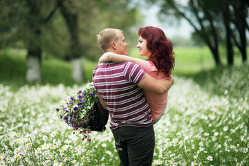 The guy holds the girl in his arms in a green park with white daisies