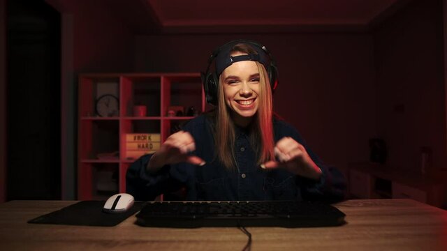 Attractive female gamer in a headset on her head plays video games at home on the computer and streams, looks at the camera with a smile on her face and shows a heart gesture.