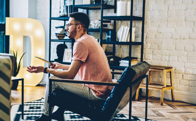 Caucasian hipster guy resting in leather chair puzzled with received email message during touch pad networking, millennial man in casual wear holding digital tablet and feeling confused in flat