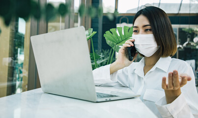 Smiling woman sitting at her desk in cafe. Happy business woman sitting in cafe and talking with present to customer by her laptop. Wear face mask for protect covid-19.