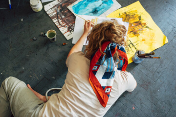 Top view of a pretty female artist sitting on the floor in the art studio and painting on paper. View from above of a woman painter with glasses painting with watercolors in the workshop.