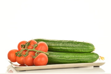 Several green cucumbers and red ripe, cocktail tomatoes on a branch with a metal tray, close-up, isolated on white.