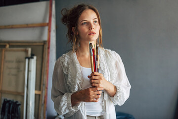 Candid portrait of an artist female posing in her art studio with paintbrushes in hands. A professional young woman painter searching for an imagination in her workshop.