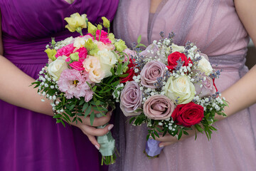 Bridesmaids or close female guests wearing beautiful tulle gowns in lavender and lilac and holding elegant mixed bouquets, close-up on the women's hands carrying flower arrangements for the wedding