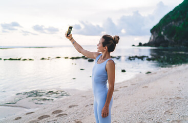 Caucasian girl with cellphone clicking selfie pictures during morning workout at seashore beach, millennial female blogger shooting influence video for sharing to social network sportive web lesson