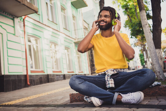 Photo portrait of guy sitting on the ground enjoying music smiling on city street in summer wearing casual outfit glasses
