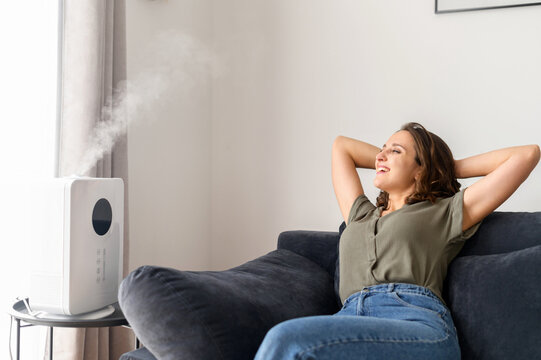 Calm and relaxed young woman enjoying fresh moisturized air from modern air humidifier sitting on the couch at home, attractive female put her hands behind her head and rests under steam