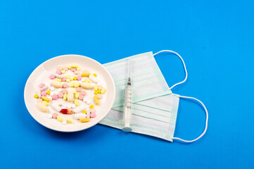 A variety of pills on a plate, medical masks and syringes next to each other on a blue background close-up.
