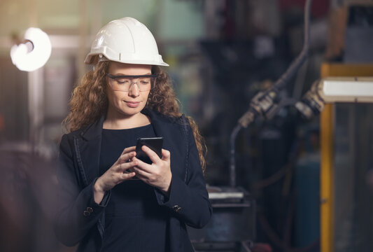 Female Industrial Worker In The Hard Hat Uses Mobile Phone While Walking Through Heavy Industry Manufacturing Factory.