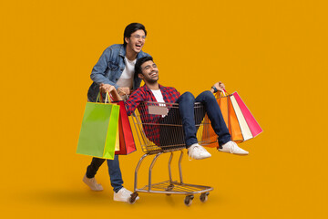 Two young men laughing with one of them sitting in a trolley holding carry bags.	