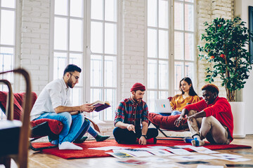 Diverse group of male and female designers cooperating on sketches sitting in flat apartment, multicultural people planning strategy of collaborative process discussing reports in home interior