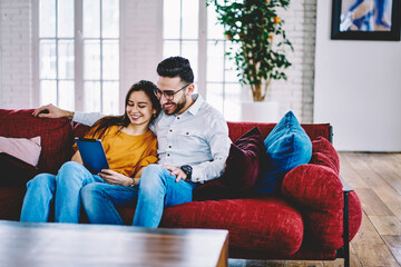 Cheerful Caucasian girlfriend and Middle Eastern boyfriend watching positive movie during weeknd pastime in home apartment, diverse couple in love enjoying relations using touch pad for networking