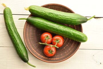 Several green cucumbers and red ripe, cocktail tomatoes on a branch with a clay plate, close-up, on a wooden white table.