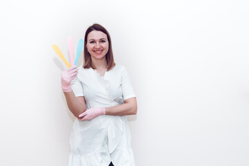 Doctor beautician in a white medical gown on a gray background holds a cosmetic spatula with place for text or logo. A woman in a white medical gown holds a white plastic cosmetic spatula.