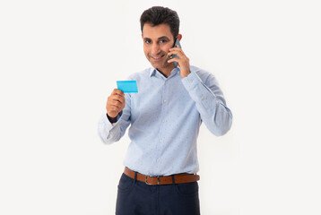 A HAPPY PROFESSIONAL LOOKING AT CAMERA WHILE HOLDING VISITING CARD AND TALKING ON MOBILE 	