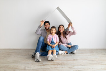 Family protection. Happy arabic family under cardboard roof sitting together on floor against white wall