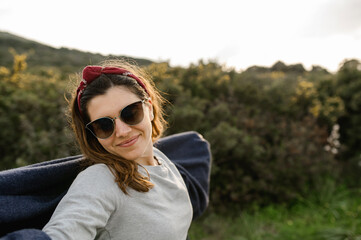 Happy casual young woman with sunglasses and scarf in countryside smiling, looking at the camera enjoying the landscape and fresh air.