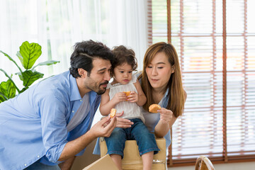 Happy young father, mother and little girl playing and eating sweet dessert together while moving into new home. Little girl eating snacks while sitting on cardboard box with father and mother at home