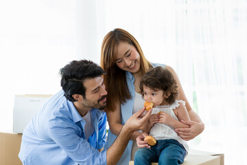 Happy young father, mother and little girl playing and eating sweet dessert together while moving into new home. Little girl eating snacks while sitting on cardboard box with father and mother at home