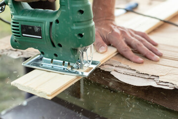 Male hands holding a jigsaw and sawing a wooden board, outdoors. 