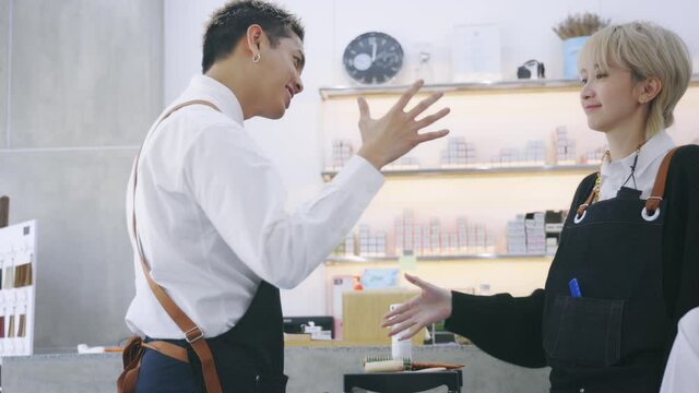 Cheerful young Asian man and woman hairdresser wearing apron uniform in modern salon giving high-five to each other on celebrating success of salon