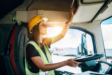 Portrait of beautiful young woman professional truck driver with protective yellow helmet sitting...