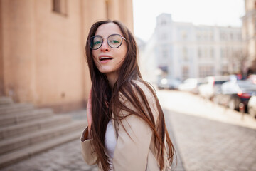 Stylish happy young woman in a jacket. Portrait of a smiling girl in glasses for sight on the street in the city center
