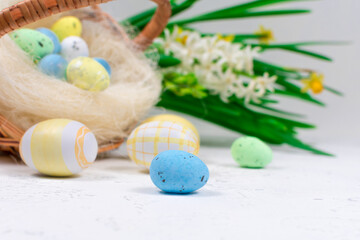 Multicolored Easter eggs in pastel colors on a white table against the background of a basket with Easter eggs and flowers of daffodils. Selective focus. Place for an inscription. Close-up.