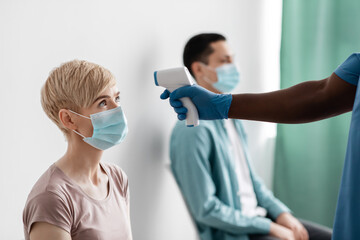 African american male nurse in protective gloves measures temperature with thermometer in adult woman in mask in clinic