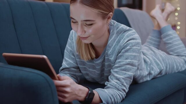 Cute young woman lying on couch using tablet computer at home evening. Relaxation, touchscreen, modern, reading, apartment, touching, convenience, paying, online, digital tablet, touchpad. Slow motion