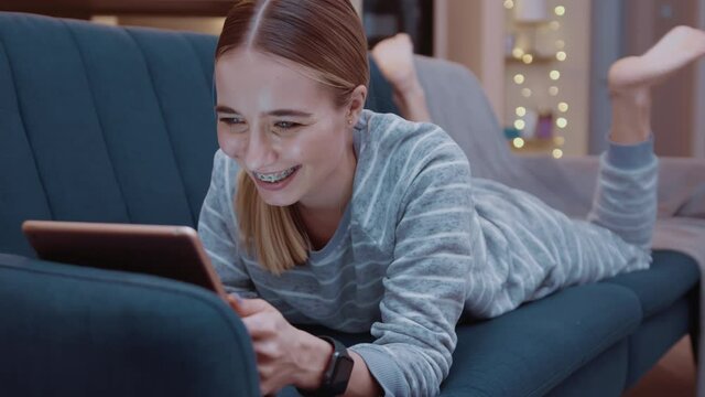 Happy young woman lying on couch using tablet computer at home evening. Relaxation, touchscreen, modern, reading, apartment, touching, convenience, paying, online, digital tablet, touchpad. Slow
