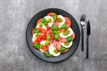 Traditional italian caprese salad with sliced tomatoes, mozzarella, basil, pesto sauce and spice on a dark background. Top view.