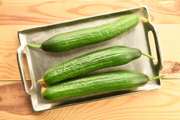 Three green cucumbers on a metal tray, close-up, on a wooden table, top view.